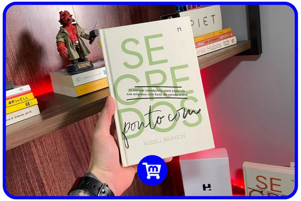 Trilogia Segredos H1 Editora: O Guia para Empreendedores em 2025 7 Livro Segredos Ponto Com de Russell Brunson em edição da H1 Editora sendo segurado em frente a uma estante.