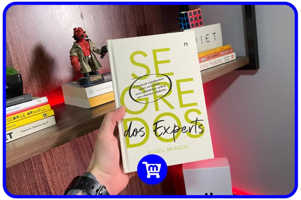 Trilogia Segredos H1 Editora: O Guia para Empreendedores em 2025 9 Livro Segredos dos Experts de Russell Brunson em edição da H1 Editora sendo segurado em frente a uma estante.