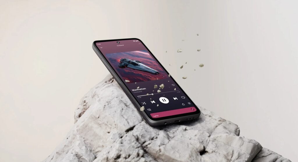 Smartphone Moto G37 apoiado sobre uma rocha clara, com a tela exibindo um aplicativo de música com capa espacial e controles de reprodução.